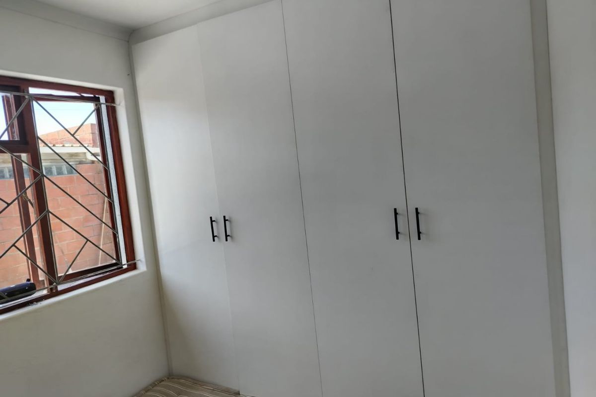 A clean bedroom featuring white built-in cupboards and a window with security bars, offering a bright and functional storage space.