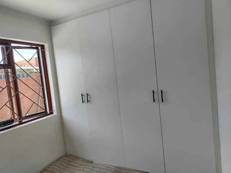 A clean bedroom featuring white built-in cupboards and a window with security bars, offering a bright and functional storage space.