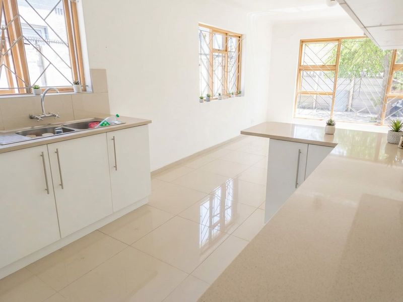 A bright, modern kitchen featuring clean white cabinetry, stone countertops, tiled flooring, and large windows with security bars.