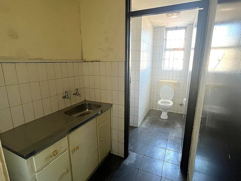 A dated bathroom and utility area featuring tiled surfaces, a stainless steel sink, built-in cabinetry, and a separate toilet area with security bars.