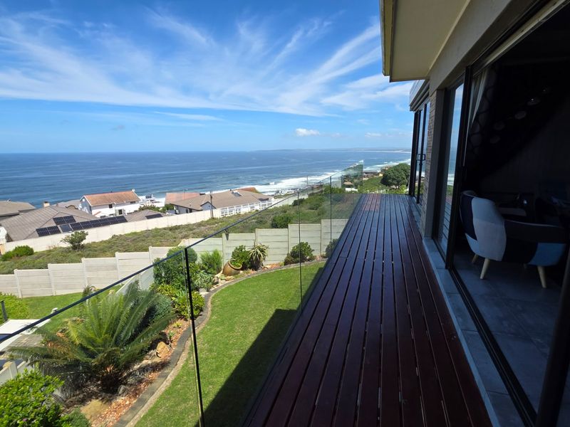 A spacious timber balcony featuring a modern frameless glass balustrade, offering expansive ocean views and direct access to the interior living space.