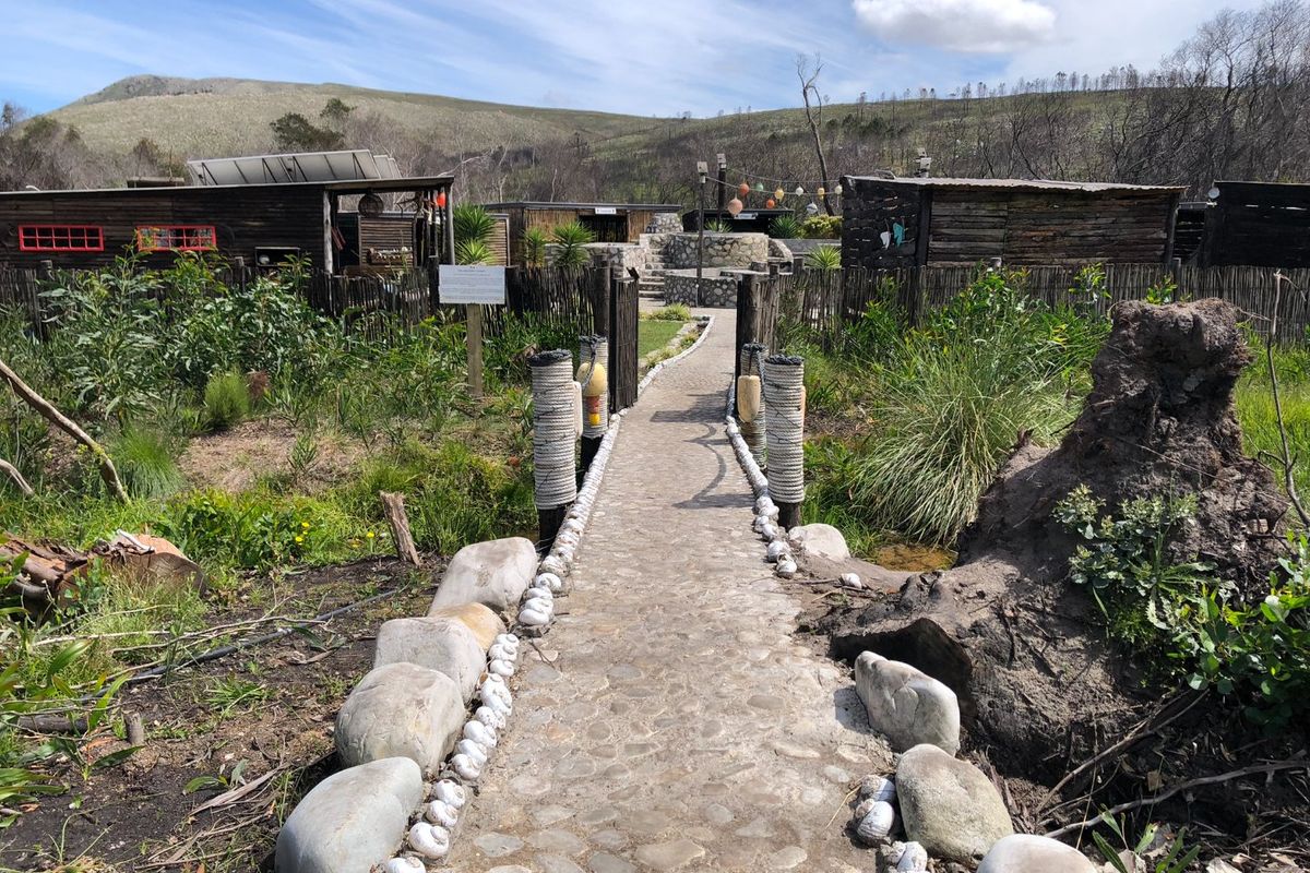 A rustic garden pathway featuring stone paving and timber fencing leading towards wooden outbuildings in a rural farm setting.