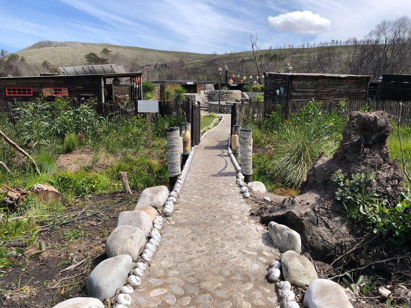 A rustic garden pathway featuring stone paving and timber fencing leading towards wooden outbuildings in a rural farm setting.