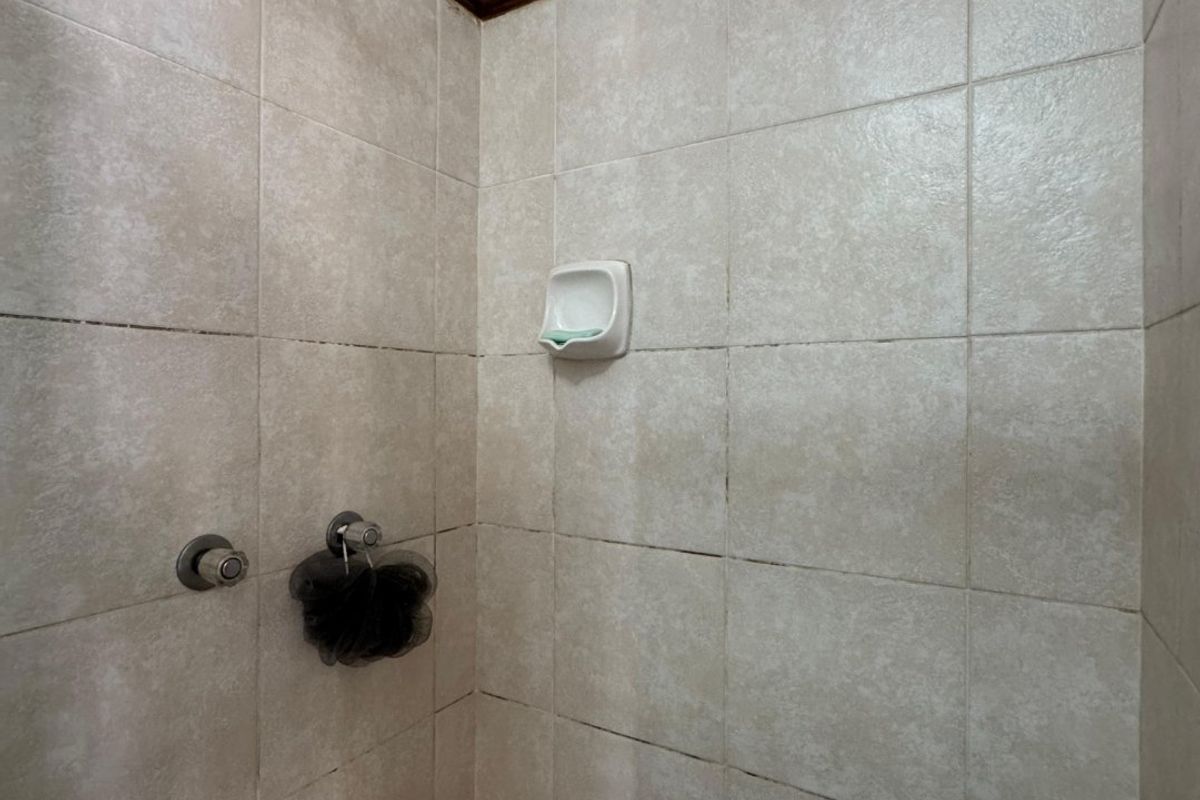 A compact shower enclosure featuring neutral wall tiles, a built-in soap dish, and standard shower fixtures, currently showing signs of wear in the grout.