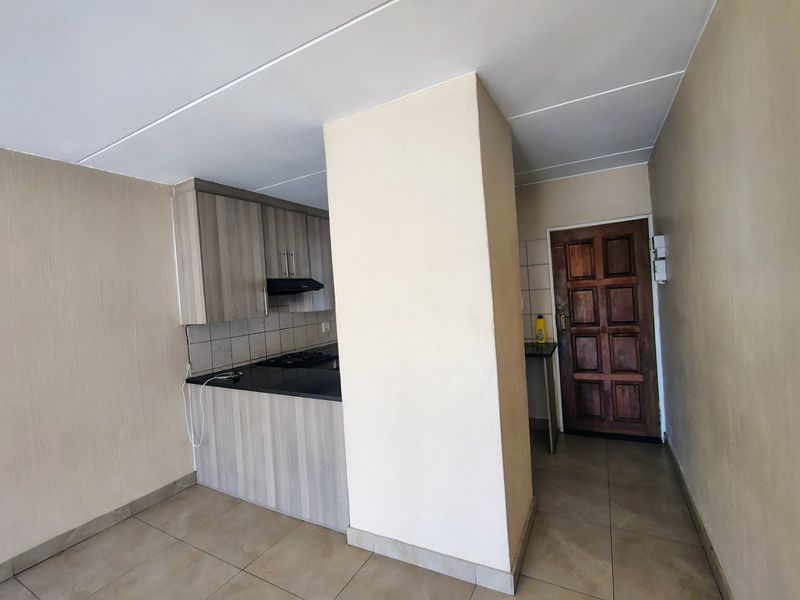 A compact kitchen area featuring built-in cabinetry, a hob with extractor fan, tiled backsplash, and neutral floor tiles, adjacent to the main entrance door.