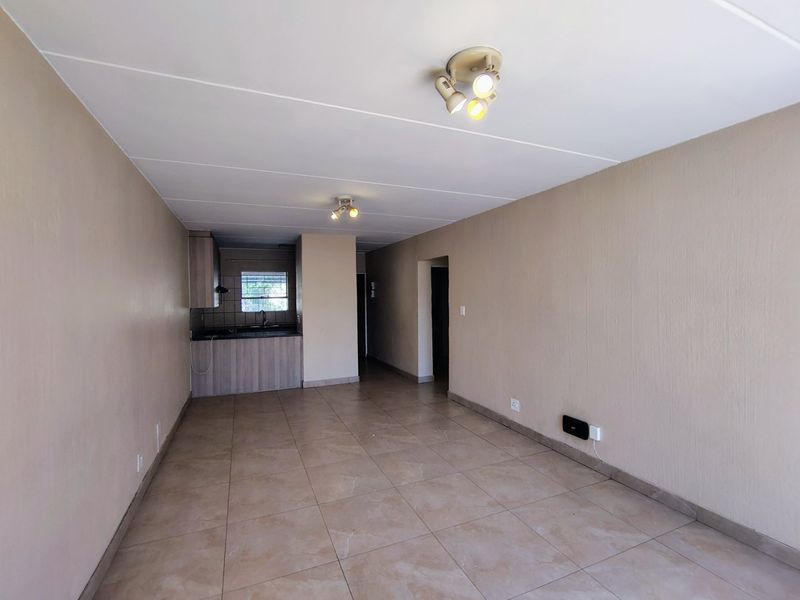 An open-plan living and kitchen area featuring neutral tiled flooring, built-in cabinetry, and functional ceiling lighting in a clean, vacant space.