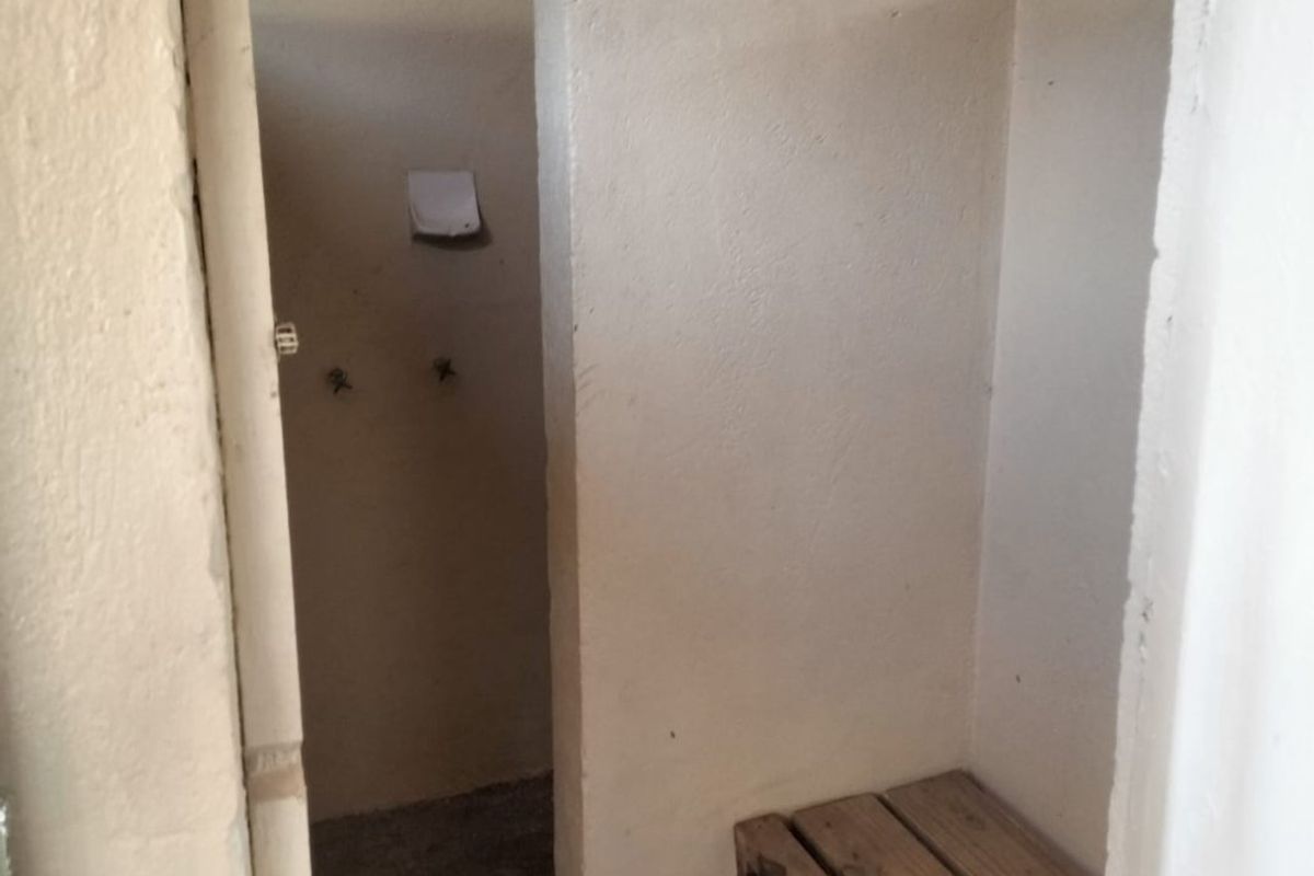 A basic, utilitarian shower area featuring exposed plumbing, concrete flooring, and a built-in wooden bench, requiring significant renovation to modernize.
