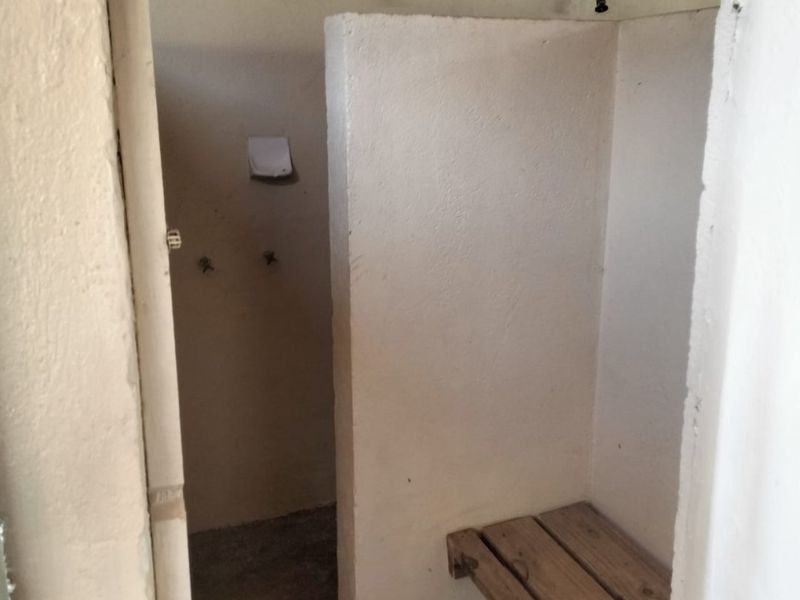 A basic, utilitarian shower area featuring exposed plumbing, concrete flooring, and a built-in wooden bench, requiring significant renovation to modernize.