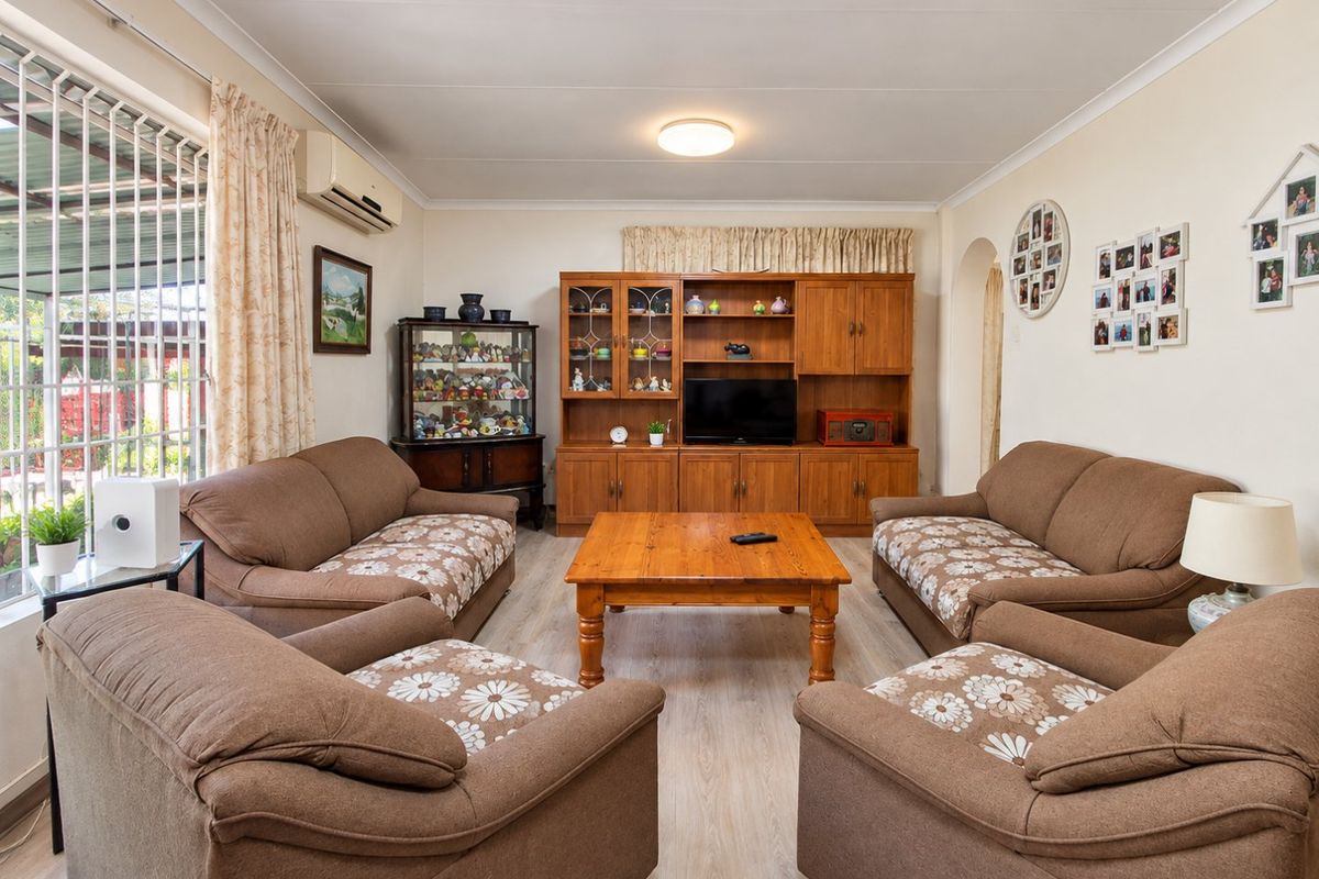 A comfortable living room featuring light-toned laminate flooring, a wall-mounted air conditioning unit, and large windows fitted with security bars.