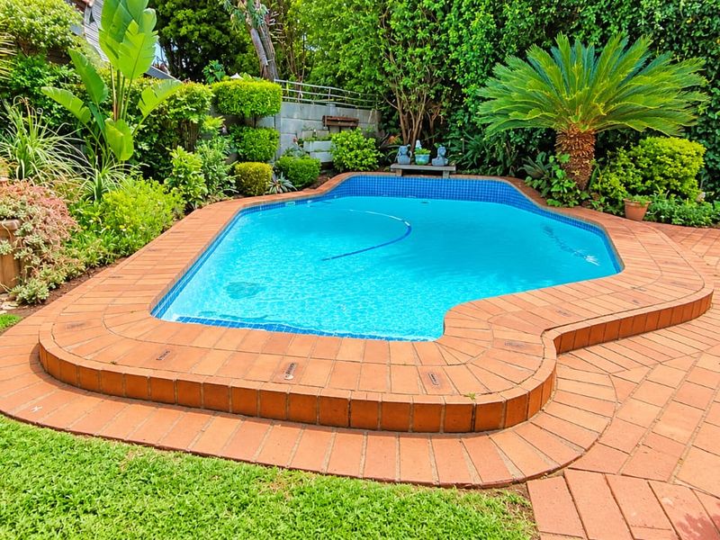 A well-maintained swimming pool area featuring brick coping, paved patio surround, and established garden landscaping.