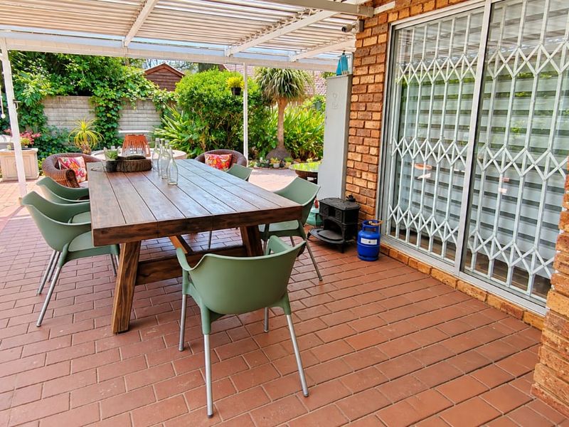 A covered outdoor patio featuring brick paving, an exposed brick wall, and a sliding glass door with a security gate, overlooking a garden.