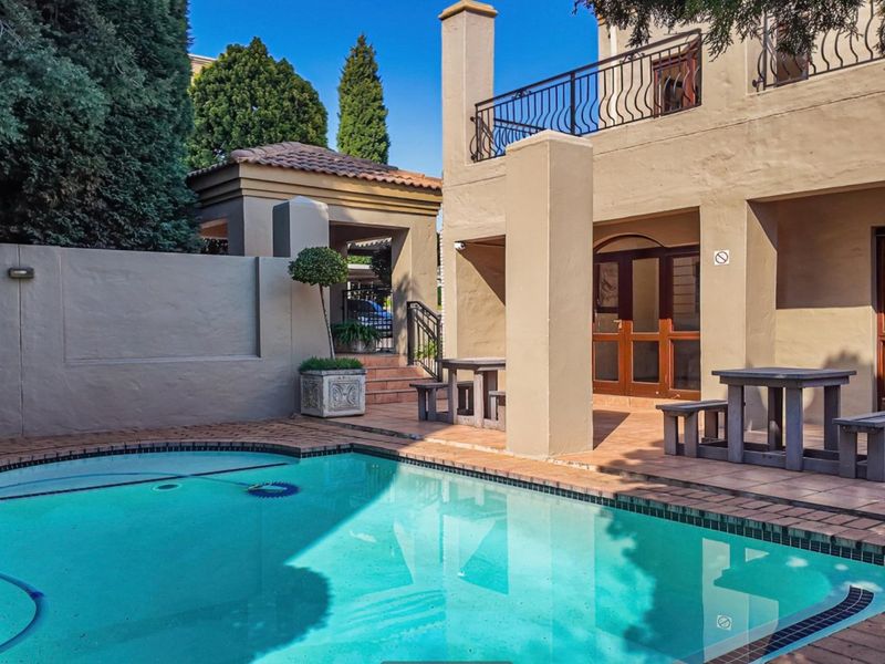 A well-maintained outdoor pool area featuring a paved patio, covered terrace space, and a swimming pool set against a masonry boundary wall.