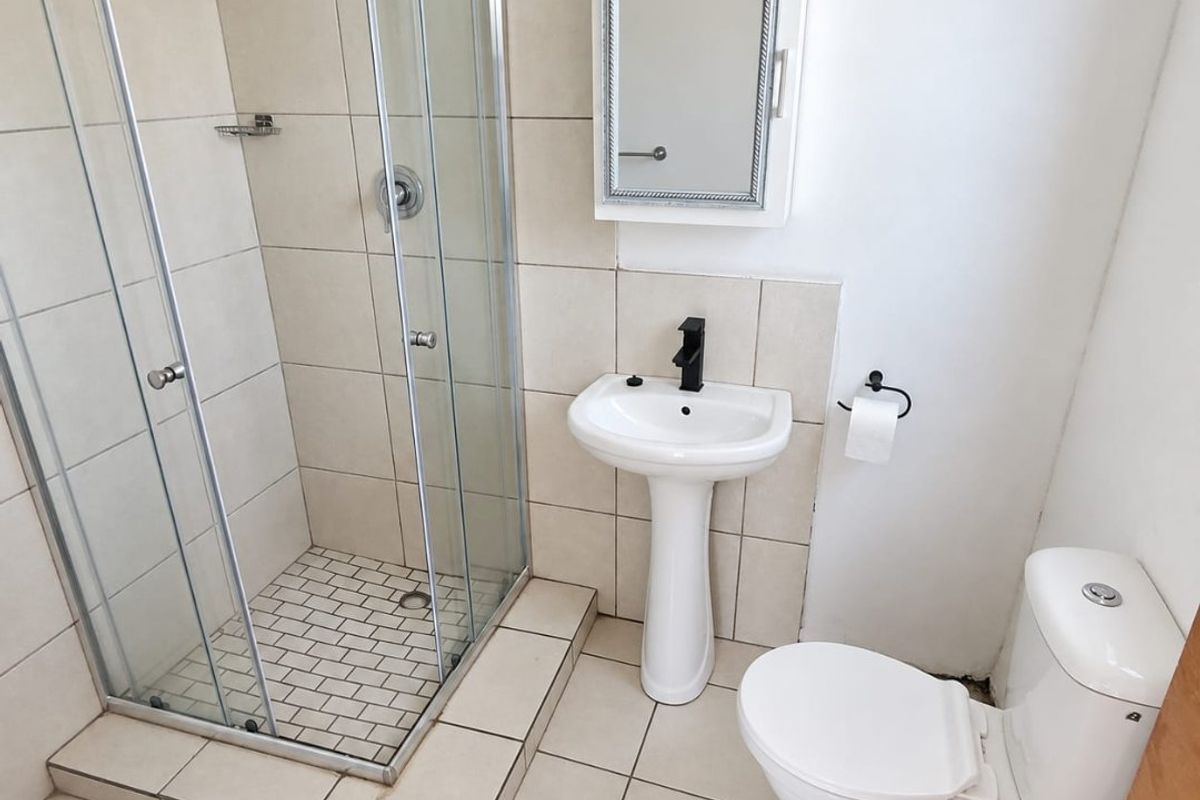 A functional bathroom featuring a glass-enclosed shower, pedestal sink, and toilet with neutral wall and floor tiling.