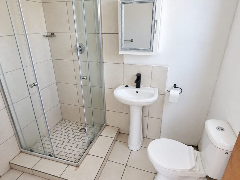 A functional bathroom featuring a glass-enclosed shower, pedestal sink, and toilet with neutral wall and floor tiling.
