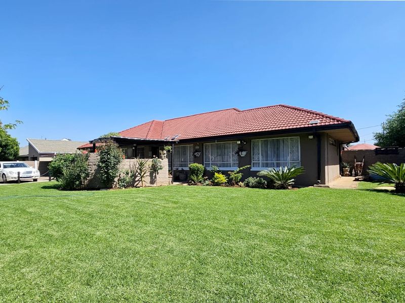 A well-maintained rear exterior view of a single-story house featuring a tiled roof, brick facade, and a spacious green lawn.