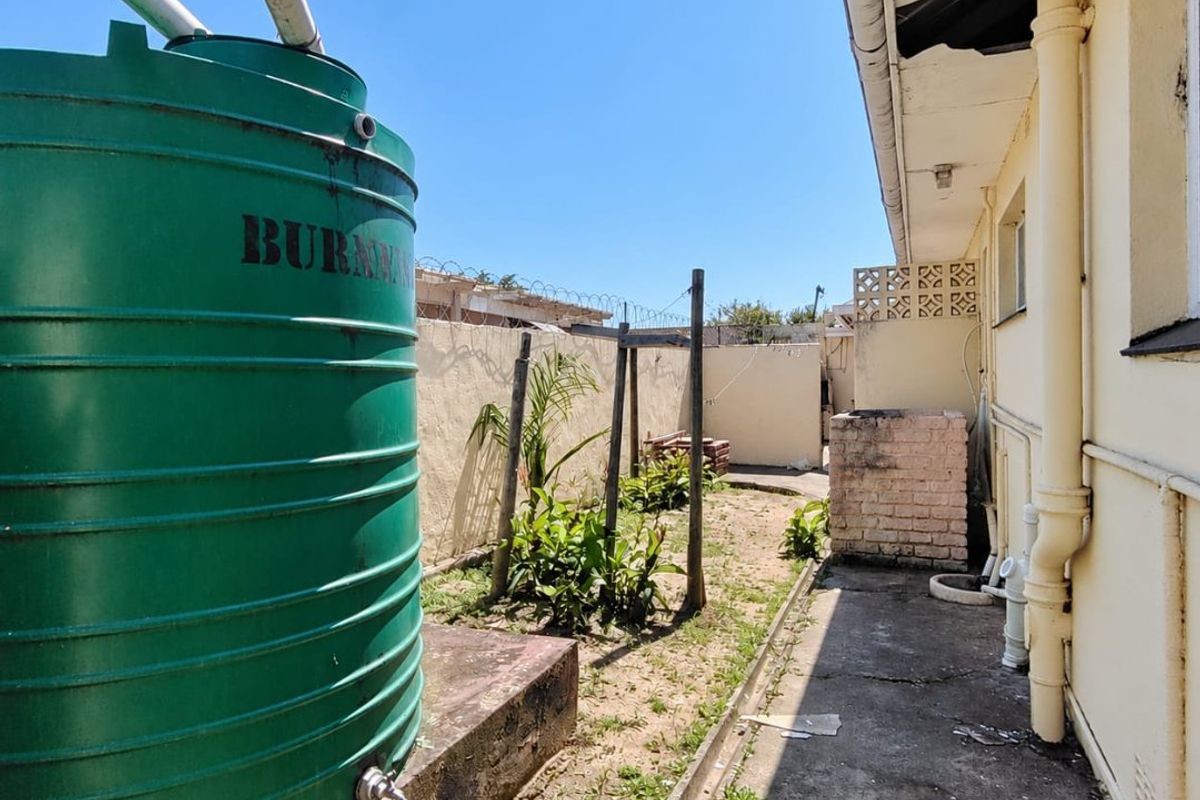 Rear exterior area featuring a large water storage tank, concrete pathway, and brick masonry wall with security fencing.