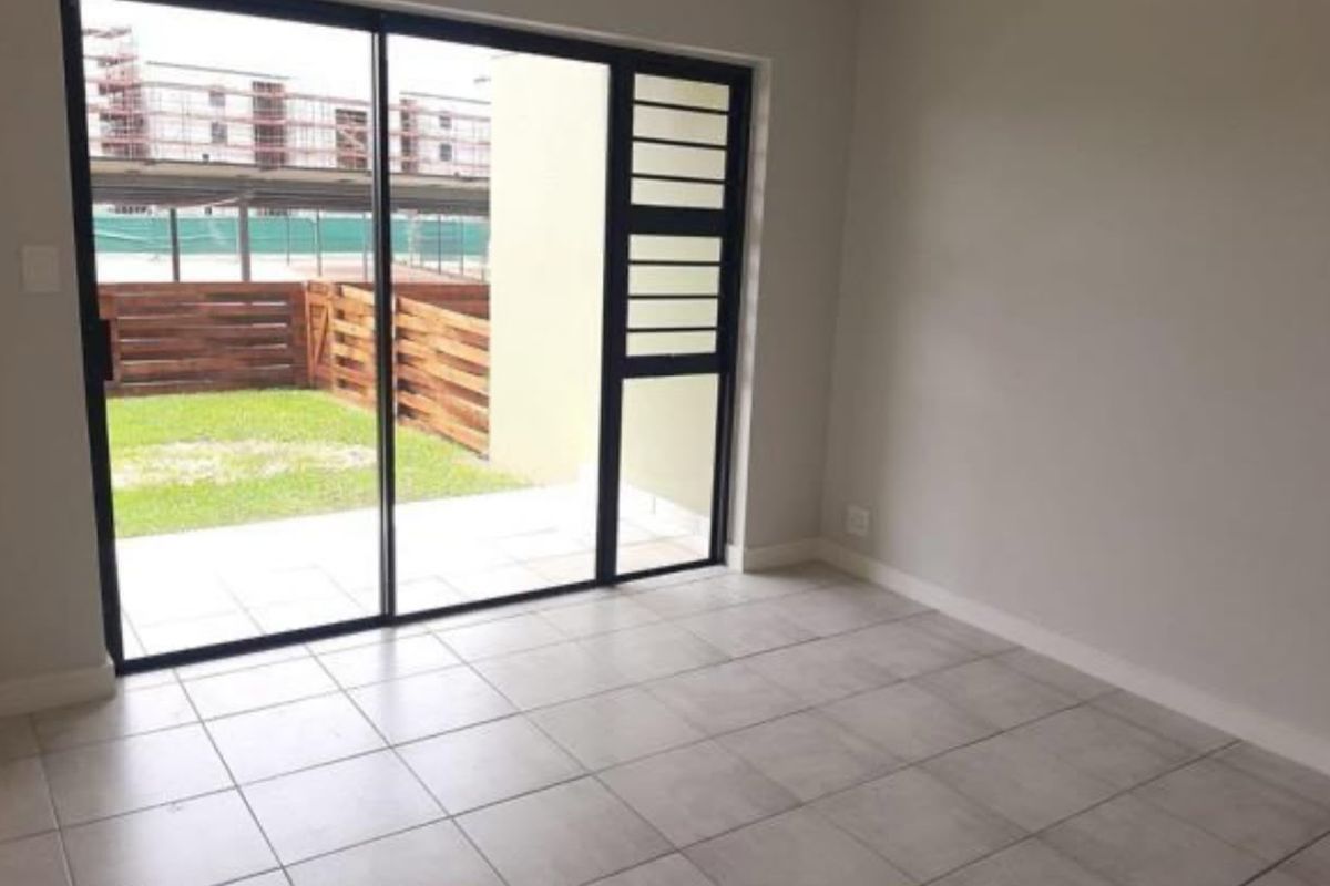 A bright, empty living area featuring neutral tiled flooring and a large sliding glass door providing direct access to a private patio and garden.