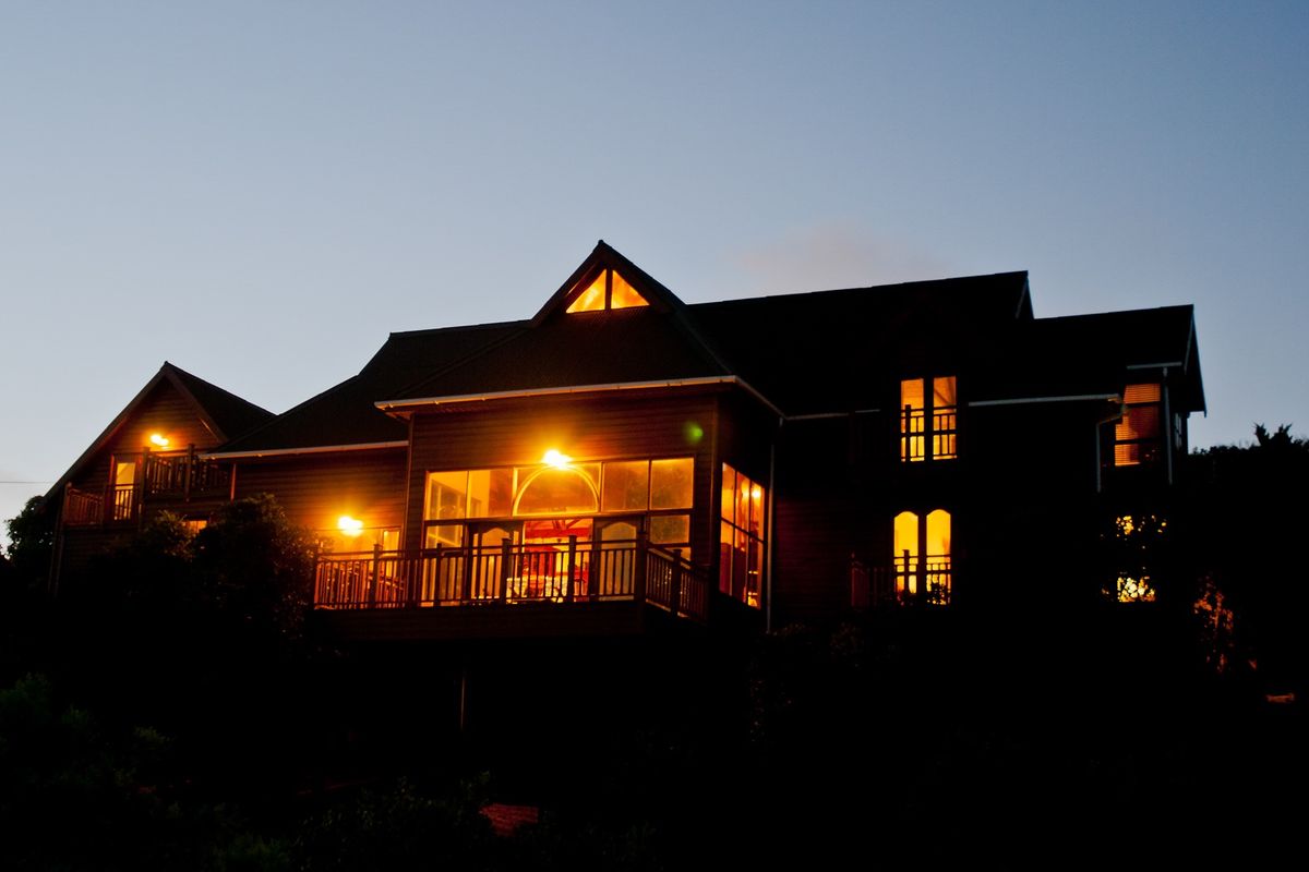 A twilight view of the rear elevation of a multi-story house featuring extensive timber decking, large windows, and a pitched roof design.