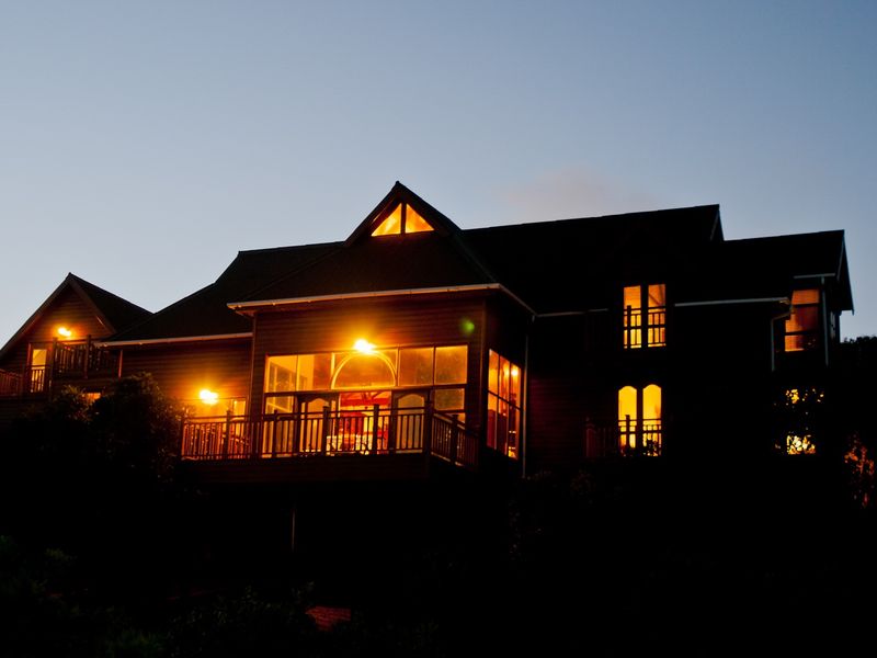 A twilight view of the rear elevation of a multi-story house featuring extensive timber decking, large windows, and a pitched roof design.