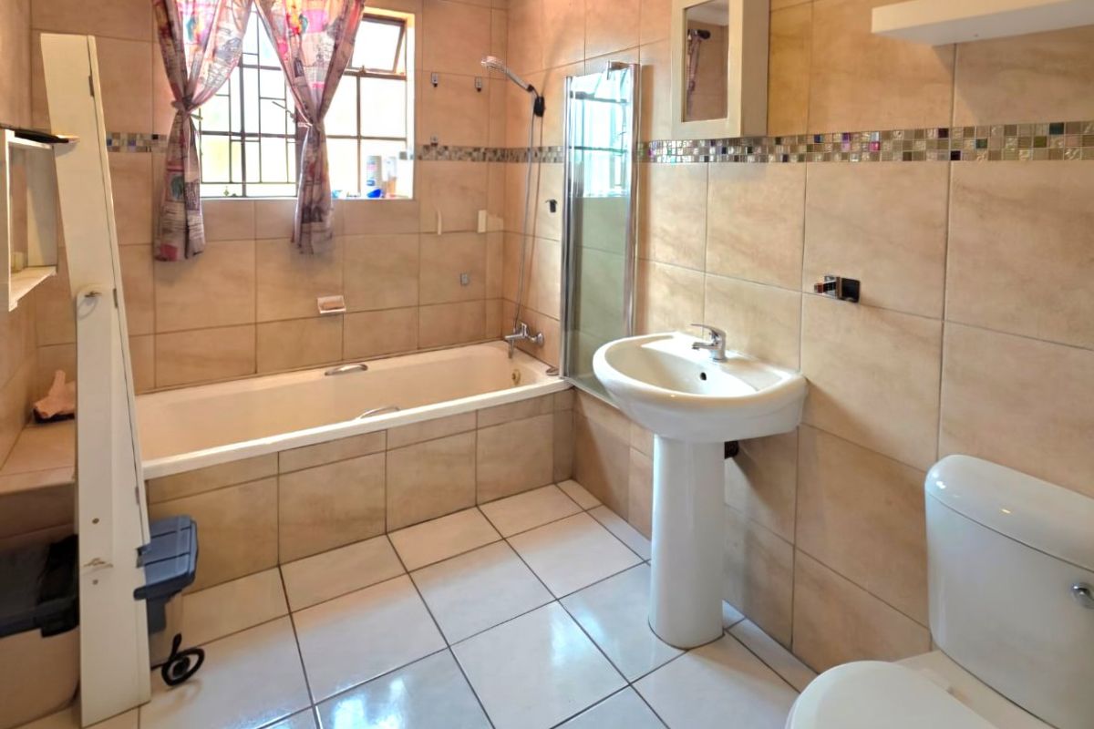 A functional bathroom featuring a bathtub with shower mixer, pedestal basin, toilet, and neutral wall and floor tiling.