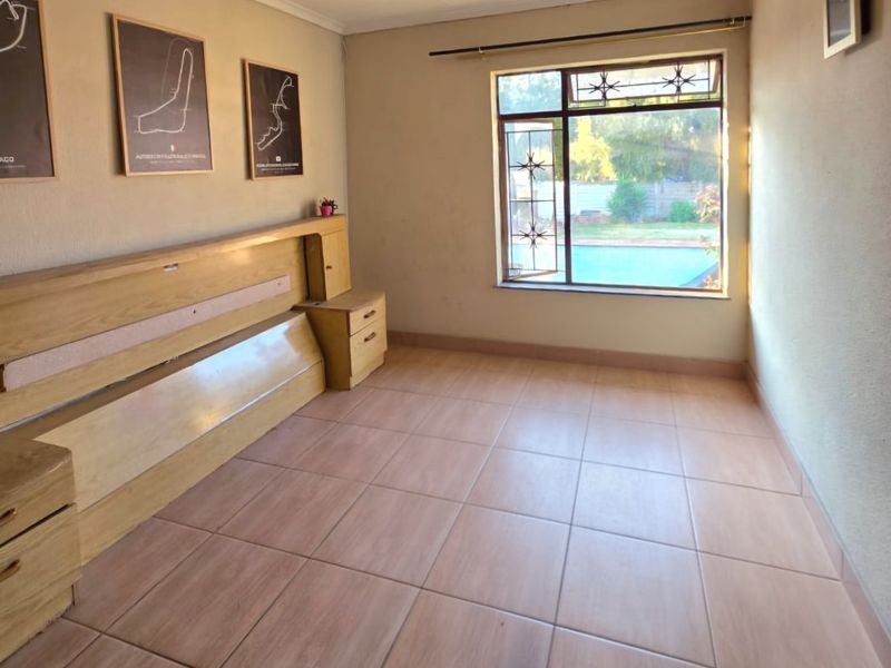 A bedroom featuring tiled flooring, a built-in headboard with integrated bedside tables, and a window with security bars overlooking the pool area.