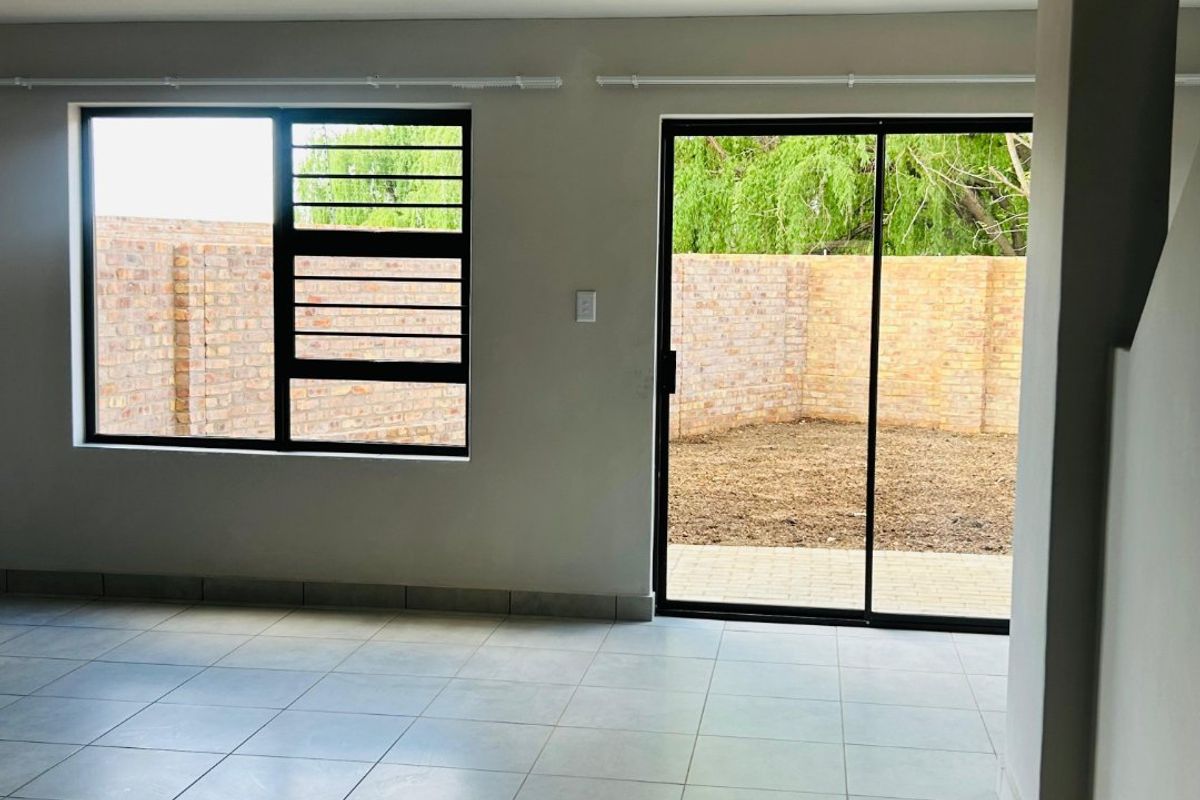 A bright, empty living area featuring neutral tiled flooring, large aluminium windows with security bars, and a sliding door leading to the rear garden.