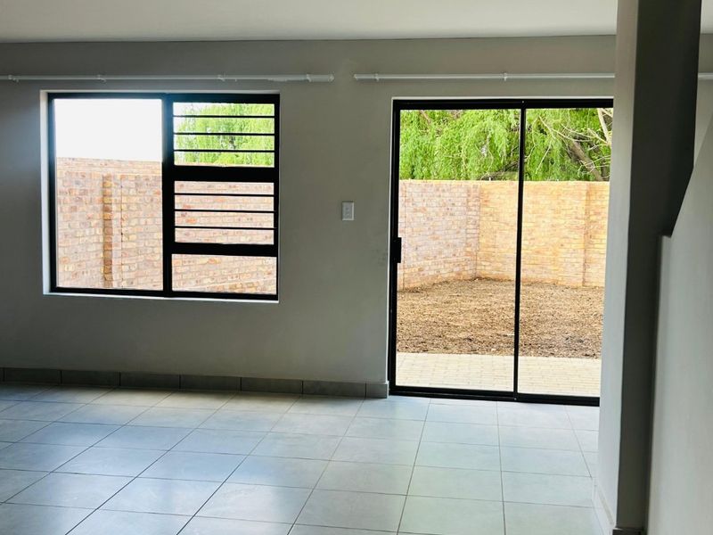 A bright, empty living area featuring neutral tiled flooring, large aluminium windows with security bars, and a sliding door leading to the rear garden.