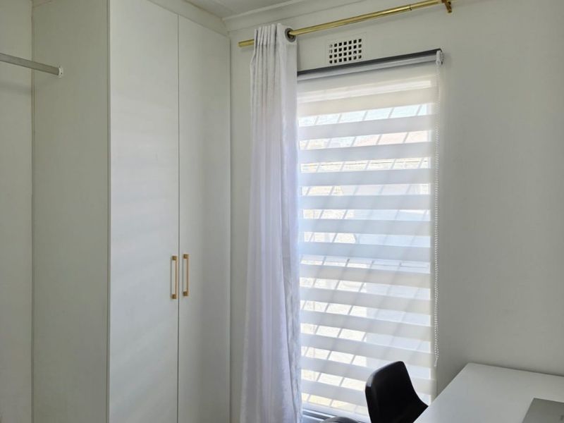 A bright bedroom featuring white built-in wardrobes, modern marble-look tiled flooring, and a window fitted with a roller blind and curtain rod.