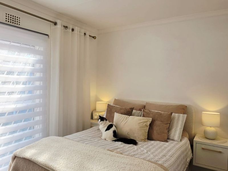 A well-maintained bedroom featuring neutral tones, a ceiling light fixture, and a window with fitted blinds and curtains.