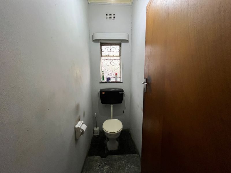 A small, dated guest toilet featuring a high-level cistern, security gate on the window, and worn wall and floor finishes.