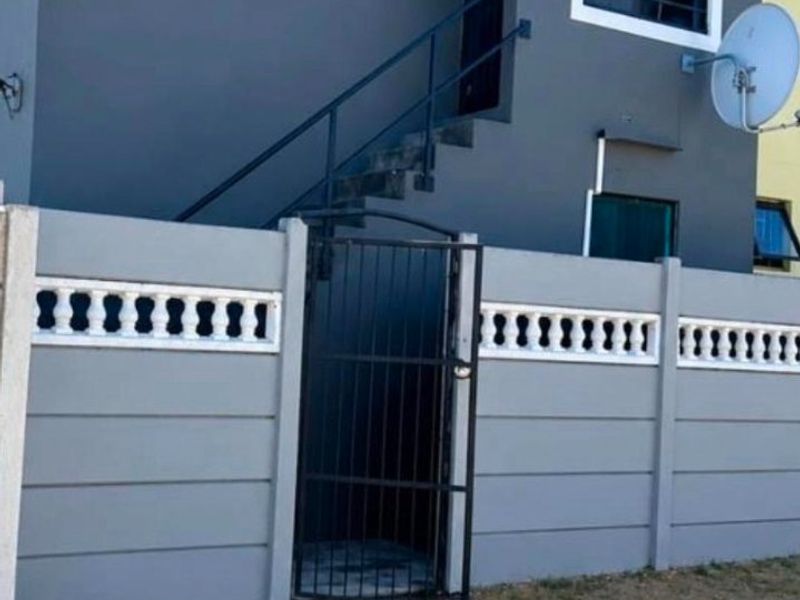 Front exterior view of a two-story residential property featuring a grey facade, external staircase, and a secure precast concrete boundary wall with a metal gate.