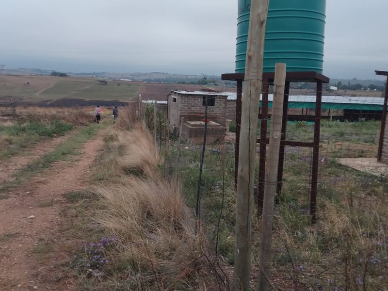 Outdoor farm area featuring a water storage tank on a metal stand, wire fencing, and a small brick outbuilding along a dirt track.