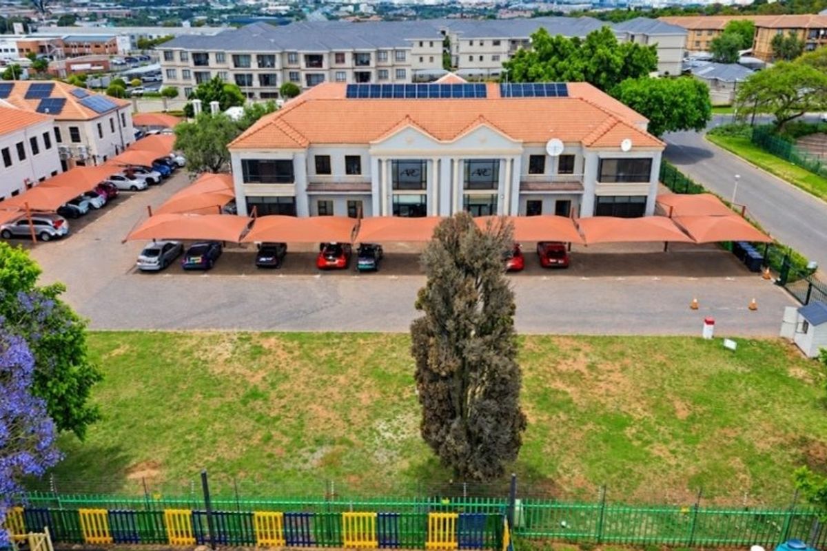 Aerial view of a commercial office complex featuring a tiled roof with solar panels, extensive paved parking with shade cloth carports, and perimeter security fencing.