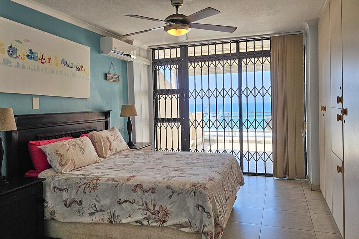 A bedroom featuring built-in cupboards, a ceiling fan, and air conditioning, with a security gate leading to a balcony offering ocean views.