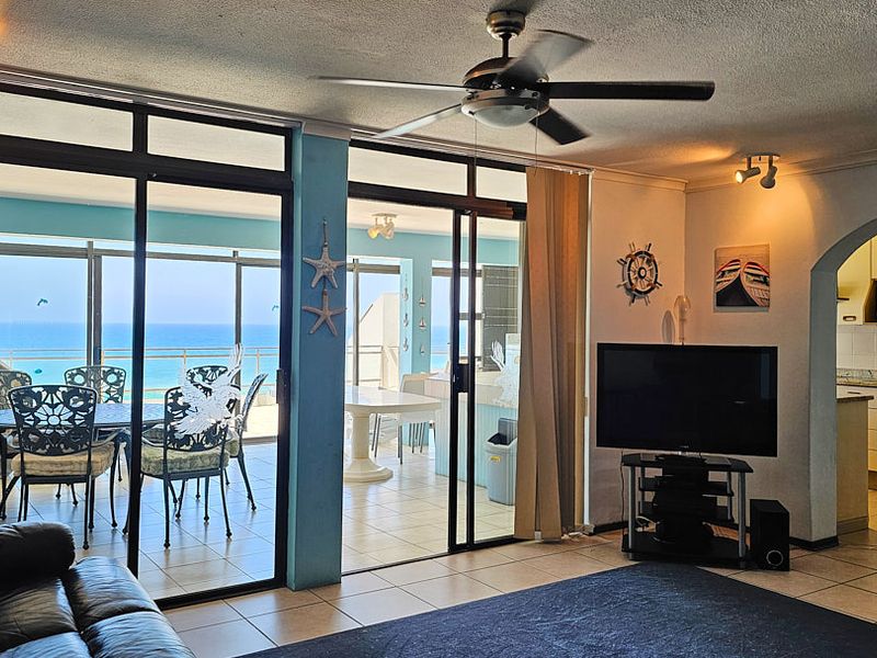 Open plan living area featuring tiled floors, a ceiling fan, and large sliding doors leading to a balcony with ocean views, adjacent to the kitchen.