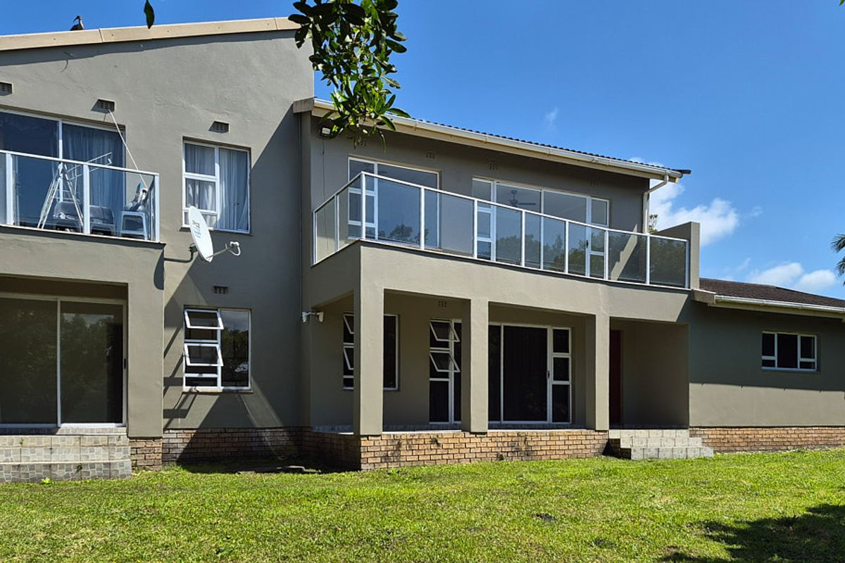 Rear elevation of a multi-story house featuring a spacious balcony with glass balustrades, sliding doors, and a well-maintained lawn area.