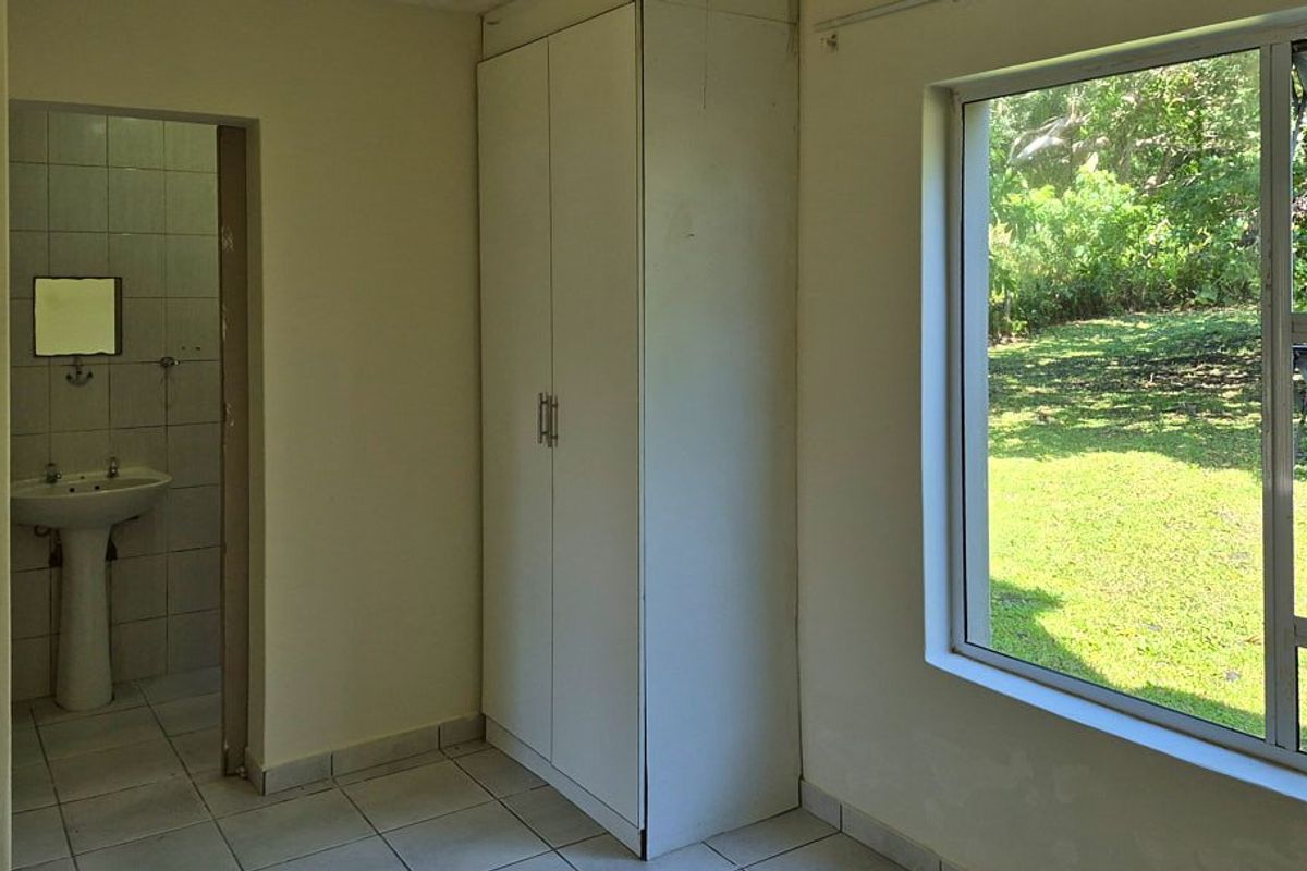 A simple bedroom featuring a built-in wardrobe, tiled flooring, and an en-suite bathroom with a pedestal basin, overlooking a garden.