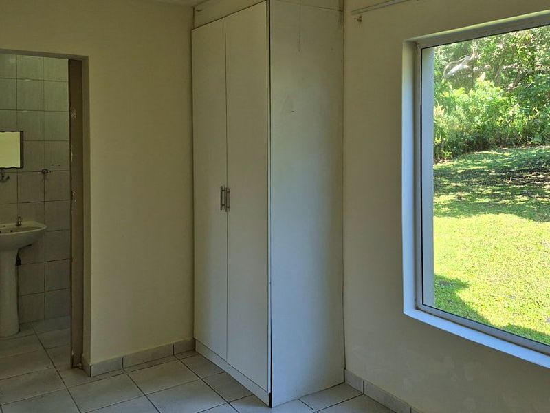 A simple bedroom featuring a built-in wardrobe, tiled flooring, and an en-suite bathroom with a pedestal basin, overlooking a garden.