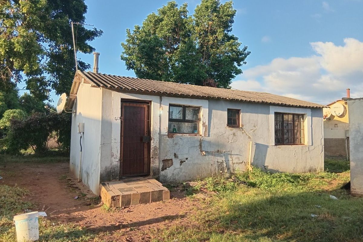 Rear view of a small detached dwelling featuring a corrugated roof, security gate, and burglar bars, showing significant wear to the exterior plaster finish.