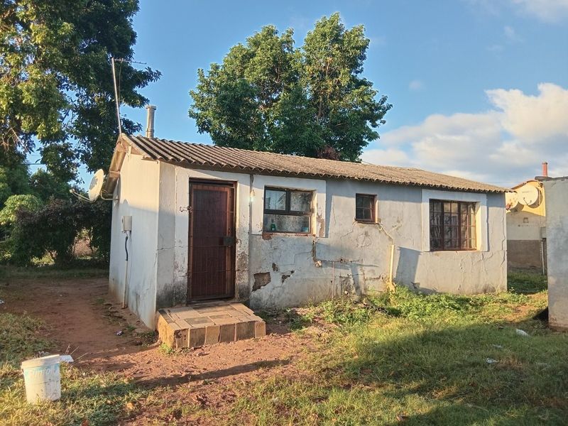 Rear view of a small detached dwelling featuring a corrugated roof, security gate, and burglar bars, showing significant wear to the exterior plaster finish.