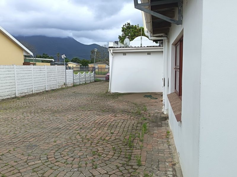 A rear exterior view showing a paved driveway area, perimeter precast walling, and a side view of the house structure with mountain views.