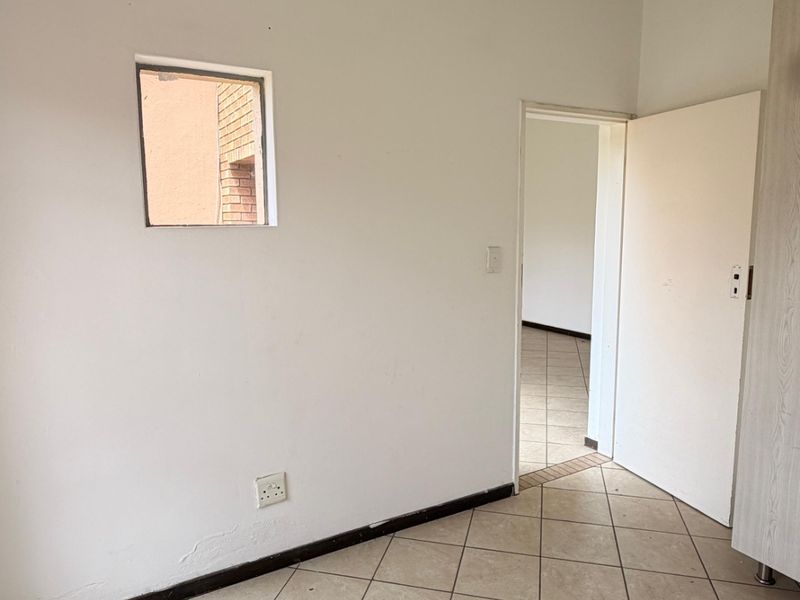 An empty bedroom featuring neutral ceramic floor tiles, a small square window, and a standard internal door, ready for renovation or staging.