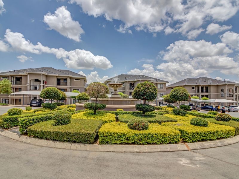 A well-maintained apartment complex entrance featuring a central water fountain, manicured landscaping, and covered parking facilities.