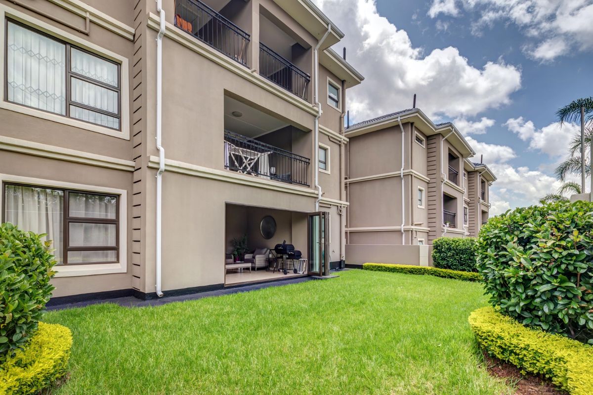 Rear view of a multi-story apartment complex featuring private balconies, ground floor patio access, and a well-maintained communal garden area.