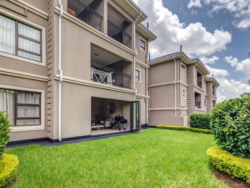 Rear view of a multi-story apartment complex featuring private balconies, ground floor patio access, and a well-maintained communal garden area.