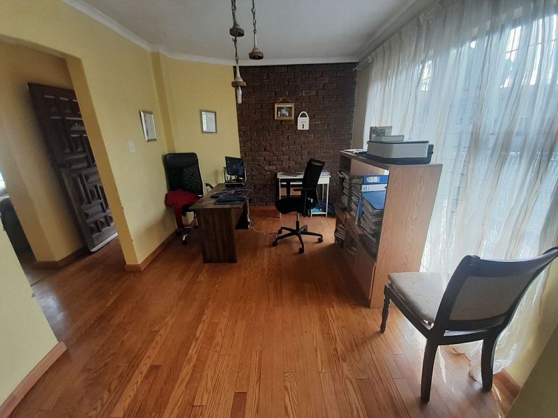 A home office featuring warm hardwood flooring, an exposed brick feature wall, and natural light through a large window.