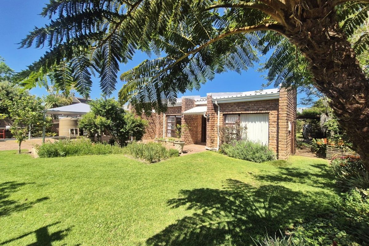 A well-maintained face brick house exterior featuring a spacious green lawn, established garden, and a paved patio area under a clear blue sky.