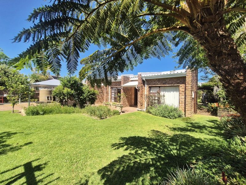 A well-maintained face brick house exterior featuring a spacious green lawn, established garden, and a paved patio area under a clear blue sky.