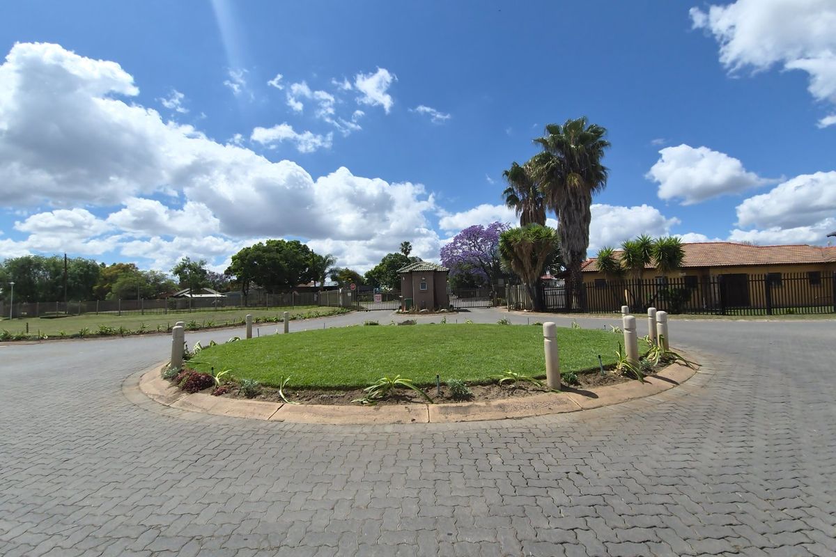 A view of a residential estate entrance featuring a paved roundabout, landscaped central island, and a security gatehouse.