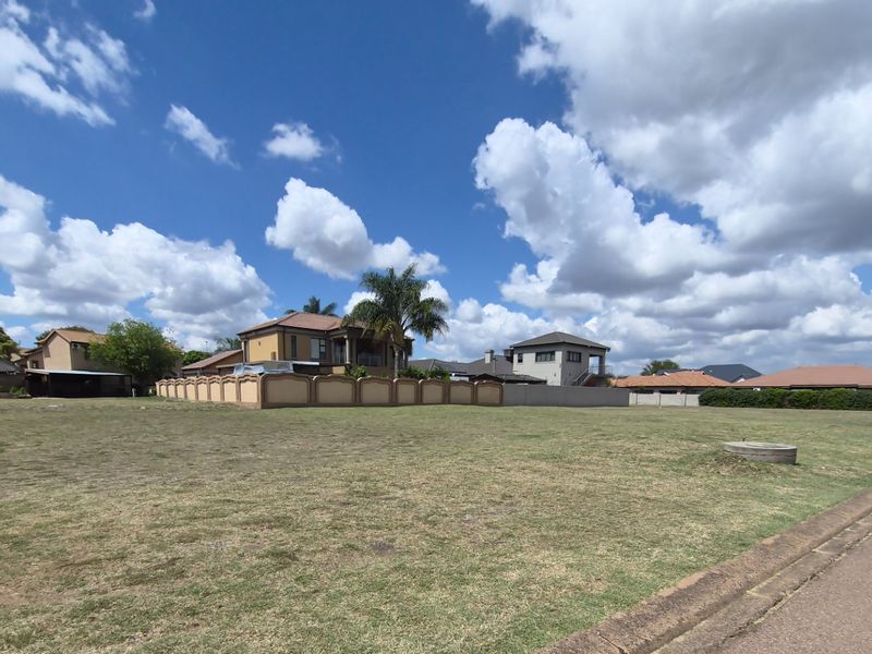 A wide-angle view of a vacant residential plot featuring a grass lawn, adjacent to a paved street and established neighborhood perimeter walls.